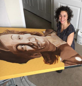 This is a photograph of artist Carrie Brummer. She is sitting on the floor of a carpeted room, with a 3 by 4 foot painting on her lap. It's a portrait of a woman and she is embroidering into her collar of her neck.