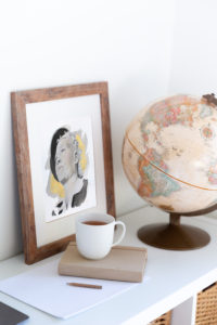Image of a bookshelf with globe, cup and book on it. There is a framed picture behind those objects leaning against the wall and has a portrait of Ruth from Carrie Brummer's Anonymous Woman series.